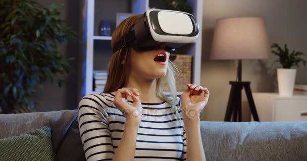 Primer plano de la joven rubia con gafas VR con auriculares VR y haciendo un gesto de miedo en el sofá en casa por la noche. Retrato. En interiores — Vídeo de stock