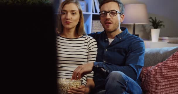 Retrato de la joven pareja caucásica riendo en voz alta, comiendo palomitas de maíz y viendo la televisión en el sofá en casa por la noche. De interior — Vídeos de Stock