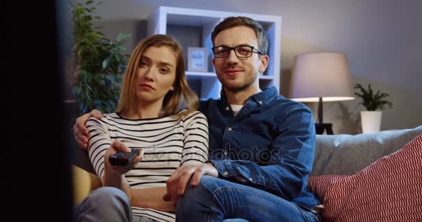 Bored young couple sitting on the sofa in the living room at night and changing TV channels with a remote control in her hand. Indoors. Portrait shot — Stock Video