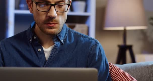 Retrato del hombre guapo caucásico en gafas que trabajan en la computadora portátil en casa por la noche. De cerca. De interior — Vídeos de Stock