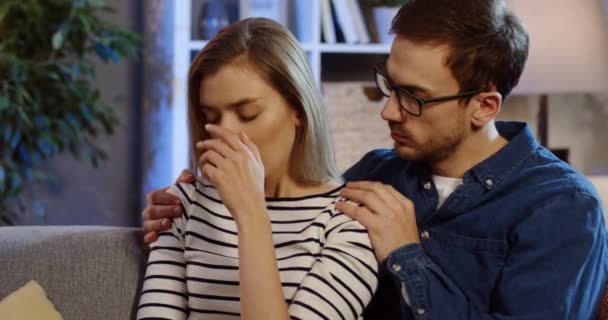 Retrato del hombre caucásico con gafas abrazando a la rubia llorando mientras se sentaban en el sofá en la habitación de casa por la noche. De cerca. De interior — Vídeos de Stock