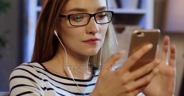 Πορτρέτο της νεαρής γυναίκας ελκυστική γυαλιά και ακουστικά taping στο smartphone και χαμογελαστή στο σαλόνι. Κοντινό πλάνο. Στο εσωτερικό — Αρχείο Βίντεο