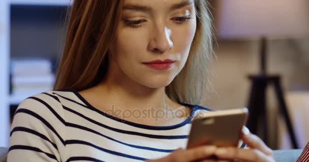 Primer plano de la joven hermosa mujer rubia escribiendo en el teléfono inteligente mientras se sienta en la noche en casa. Retrato. En interiores — Vídeo de stock