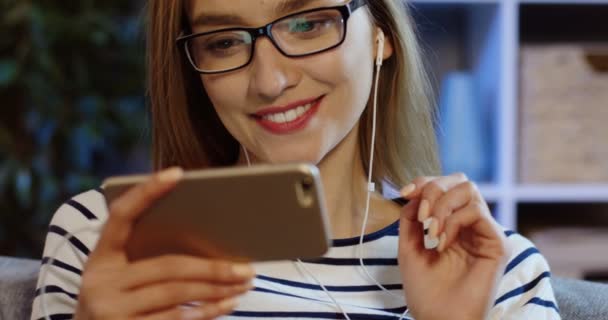 Jovem loira encantadora mulher em óculos e fones de ouvido assistindo algo no smartphone e rindo na sala de estar. Fecha. Retrato. Interior — Vídeo de Stock