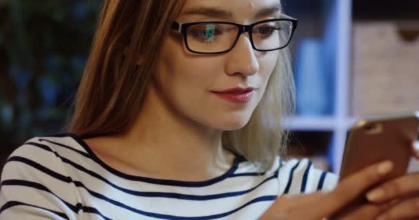 Primer plano de la joven atractiva mujer en gafas charlando en el smartphone y sonriendo en casa. Retrato. De interior — Vídeos de Stock