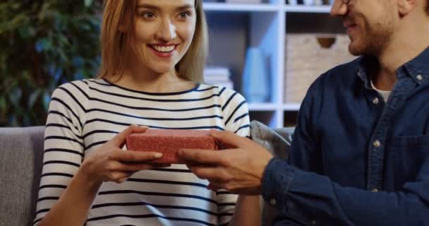 Beau homme donnant un cadeau enveloppé à sa belle petite amie et elle se demandant ce que c'est que ça. Dans le salon sur le canapé. Prise de vue portrait. À l'intérieur — Video