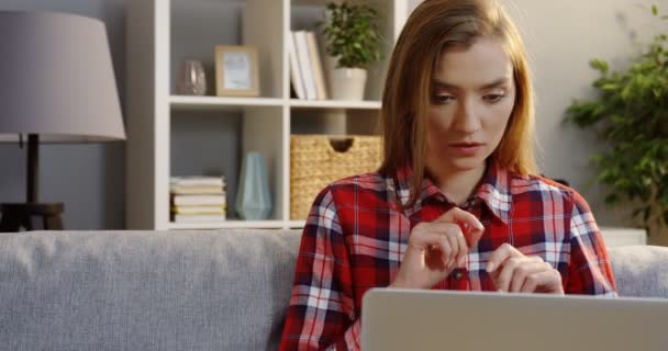 Portraitaufnahme der jungen blonden Frau im karierten Hemd, die am Laptop arbeitet, während sie auf dem Sofa im Wohnzimmer sitzt. innen — Stockvideo