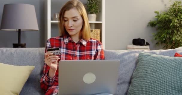 Porträtt skott av den unga vackra kvinnan i en brokig skjorta att köpa online med ett kreditkort i handen på den bärbara datorn medan du sitter i soffan i mysiga rummet. Inomhus — Stockvideo