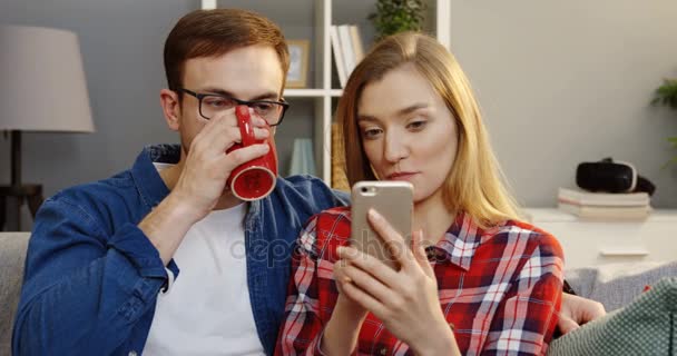 Ritratto della donna bionda affascinante che mostra qualcosa sullo schermo del suo smartphone al marito che beve il tè mentre si siede vicino a casa. Chiudete. Interni — Video Stock