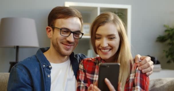 Портретный снимок молодой улыбающейся красивой женщины и мужчины, смотрящих что-то смешное на смартфоне. В помещении — стоковое видео