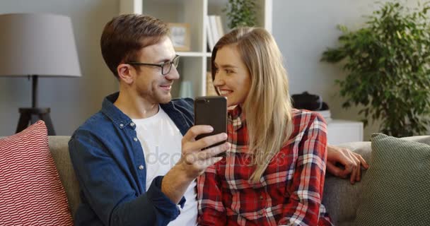 Jeune homme caucasien montrant quelque chose sur le téléphone intelligent noir à sa jolie petite amie alors qu'ils sont tous les deux assis sur le canapé dans la belle chambre. À l'intérieur — Video
