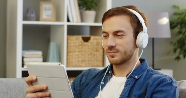 Retrato del hombre atractivo caucásico en los auriculares mirando la pantalla del dispositivo de la tableta y sonriendo en la acogedora habitación. De cerca. En interiores — Vídeos de Stock