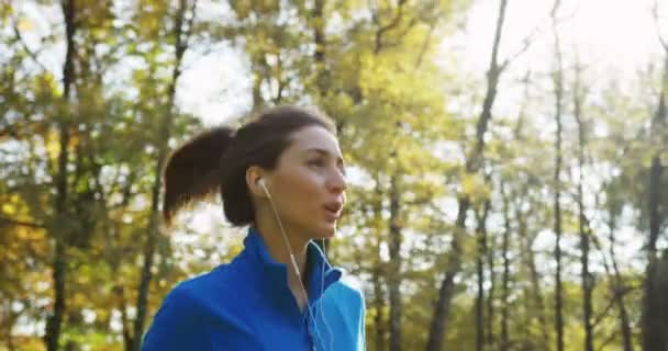 Близько з боку молодої спортивної красивої жінки в синьому светрі і з навушниками, що бігають в парку. Сонячна погода ранньої осені. На відкритому повітрі. Повільний рух — стокове відео