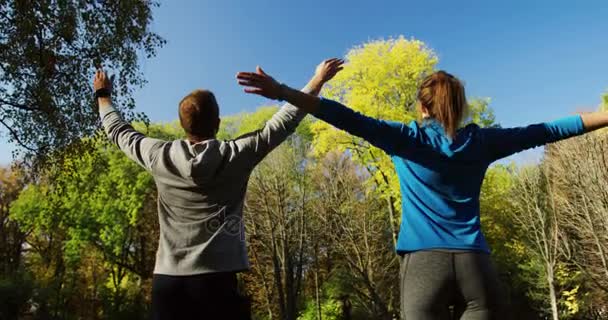 Mladá žena a muž dělá jógu na vzduch v sunny park v časných ranních hodinách. Procvičení a protažení. Zadní. Vně — Stock video