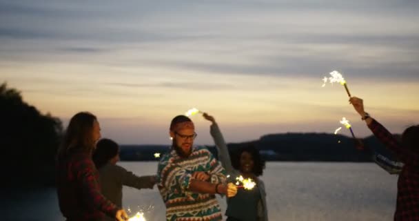 Bullriga företaget av blandade raser hipsters att ha på sjön kusten med tomtebloss pinnar i händerna i solnedgången. Utsidan — Stockvideo