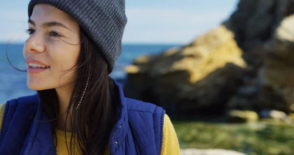 Close up of beautiful young woman in hat looking on the side and then turning her head to the camera on the rocky sea background. Cold sunny and windy day. Portrait shot. Outdoors