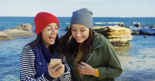 Zwei schöne junge Frauen mit Hüten, die über etwas lachen, das sie auf dem Smartphone am felsigen Ufer des Meeres beobachten. kalter, aber sonniger Tag. Außen — Stockvideo