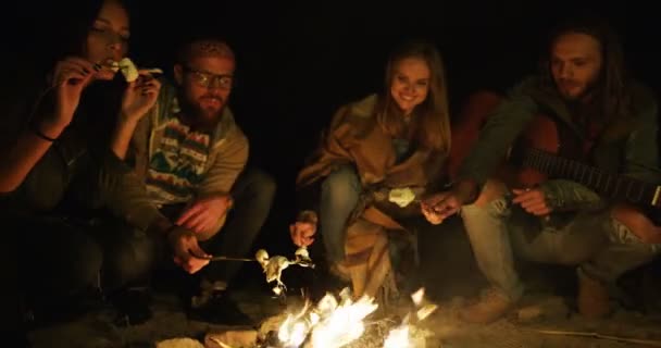 Quatre amis mâles et femelles hipsters font cuire des marshmellows sur le feu de joie et les mangent la nuit. À l'extérieur — Video