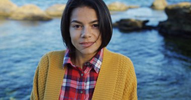 Tarafından bakarım ve kayalık deniz arka plan üzerinde kameraya gülümseyen sevimli genç kadın. Soğuk ama güneşli bir gün. Portre vurdu. Yakın çekim. Dışarıda