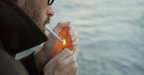 El primer plano de un hombre con gafas con barba enciende un cigarrillo en el fondo del mar. Afuera. Retrato plano —  Fotos de Stock