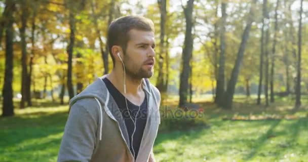 Slow motion av ung sportig kaukasiska man i hörlurar jogging i den gröna parken. Det soliga vädret i början av hösten. Utomhus. Porträtt skott — Stockvideo