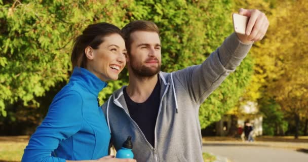 Jeune couple de coureurs souriants vidéoconférence sur le téléphone intelligent dans le parc ensoleillé au début de l'automne. À l'extérieur . — Video