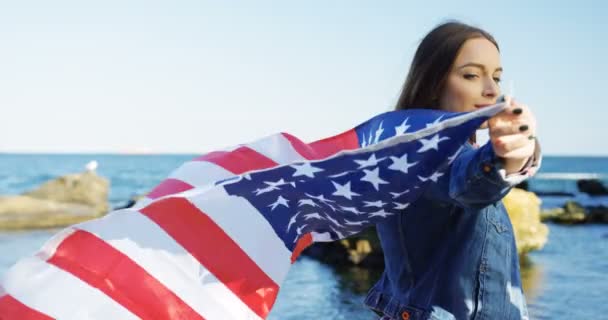 Joven mujer bonita de pie en la orilla del mar y sosteniendo una bandera americana ondeando. Disparo por la espalda y el costado — Vídeos de Stock