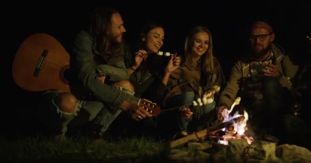 Gezelschap van stijlvolle hipster vrienden zitten rond het vuur met een gitaar en bakken marshmellows in de late avond. Buiten — Stockvideo