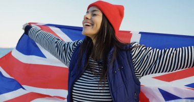 Kırmızılı kadın çekici genç bir deniz sahilinde ayakta, kameraya doğru gülen ve büyük bir İngiliz bayrağı sallayarak şapka. Mavi gökyüzü arka plan