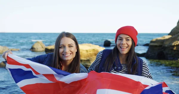 Portrait of two beautiful young women waving with the British flag and smiling on the rocky sea background. Cold but sunny day. Outside — Stock Photo, Image