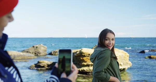 Жінка робить гарні фотографії на своєму мобільному телефоні своєї привабливої подруги, посміхаючись і даючи великі пальці на скелях і морському фоні. Холодна, але сонячна погода. На відкритому повітрі — стокове фото