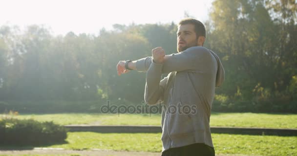 Joven hombre caucásico guapo estirando los brazos y haciendo ejercicio temprano en la mañana en el hermoso parque verde. Al aire libre — Vídeos de Stock