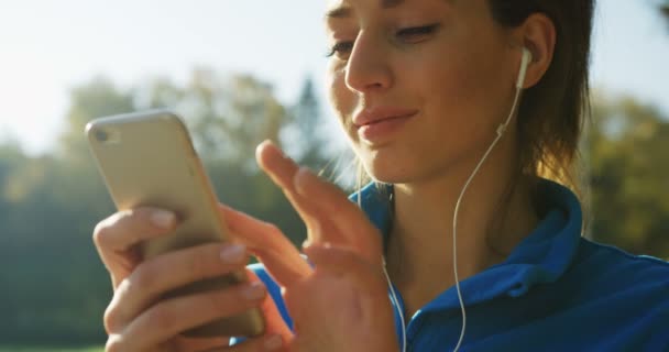 Nahaufnahme einer attraktiven Frau mit Kopfhörern, die sich im Park entspannt, während sie vor dem Laufen die Musik am Telefon auswählt und hört. sonniges Wetter. Draußen. Porträtaufnahme — Stockvideo