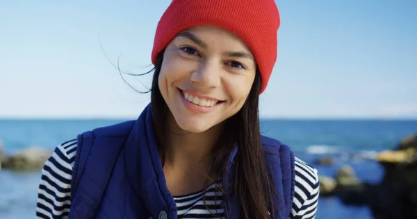 Close up of attractive young woman in red hat looking on the side and then smiling straight into the camera on the rocky sea background. Cold sunny and windy day. Portrait shot. Outdoor