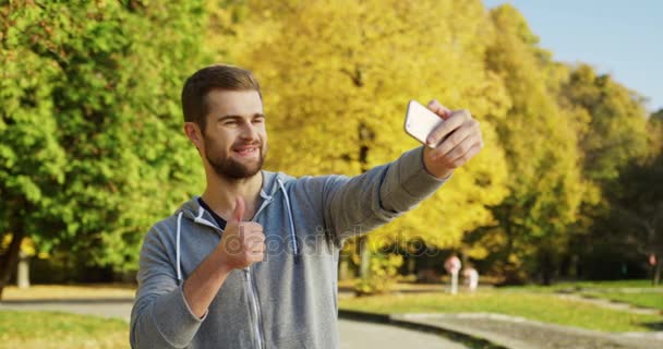Selfies をとり、秋には早い素敵な公園でスマート フォンを使用して彼の thomb をあきらめる若い白人男。外 — ストック動画