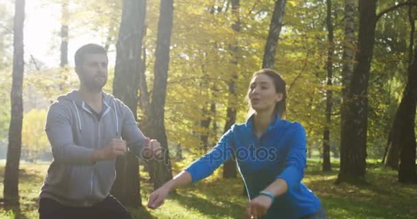 Jovem casal desportivo exercitando e agachamento no parque verde ensolarado no início da manhã. Ao ar livre — Vídeo de Stock