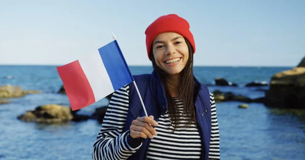 Junge schöne Frau winkt am felsigen Meeresufer mit einer kleinen französischen Flagge und lächelt aufrichtig in die Kamera. Draußen. sonniges Wetter — Stockfoto