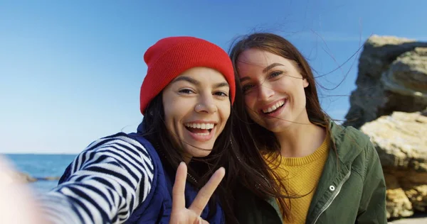 Zwei attraktive junge Frauen machen lächelnde Selfies mit Siegeszeichen auf dem felsigen Meeresgrund. kaltes, sonniges Wetter. Draußen. Das ist nicht der Fall. Nahaufnahme — Stockfoto