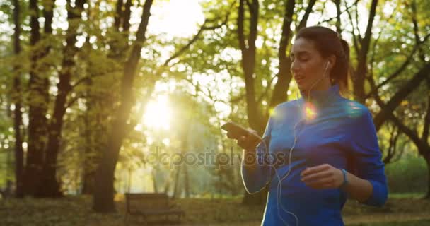 Attraktiv kvinna jogger i hörlurar stoppa i parken ändra musik på telefonen. Soligt väder. Utsidan. — Stockvideo