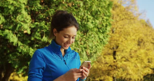 Attraktive, sportliche junge Läuferin, die vor dem Joggen im herbstlichen Park mit ihrem Smartphone klebt und lächelt. sonniges schönes Wetter. Draußen. Porträtaufnahme — Stockvideo