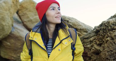 Kırmızı şapka ve kayaların arasında yürüyüş ve etrafa sarı ceket çekici genç kadın. Soğuk güneşli gün. Açık