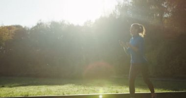 Parkta koşu kulaklık sportif kadın silueti taraftan ağır çekim. Erken sonbahar sabahı güneşli havalarda. Açık.