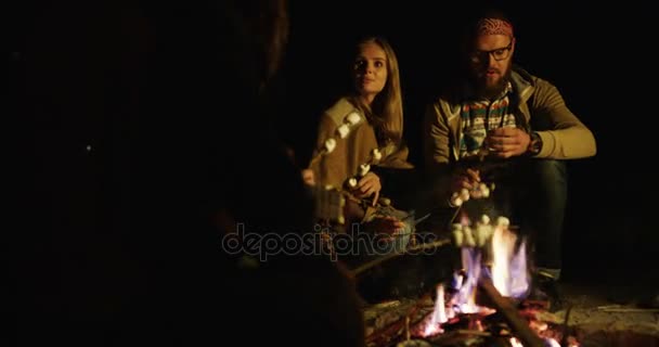 Group of hipsters friends, singing, playing the guitar, baking marshmellows on the bonfire and eating them in the night time. Outside — Stock Video