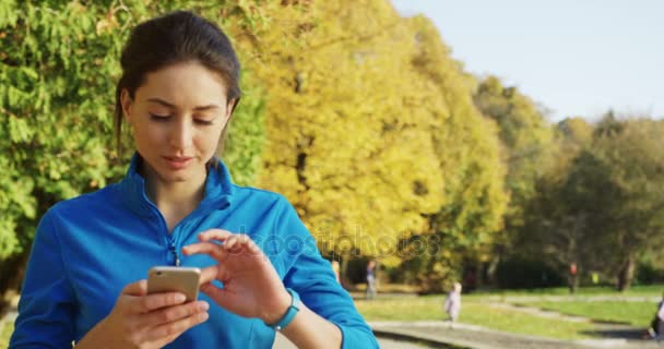 Atraktivní mladá žena v modrém svetru drží její telefon a tejpování pak po spuštění při pohledu na hodinky v parku. Pěkné slunečné počasí. Venku. Portrét — Stock video