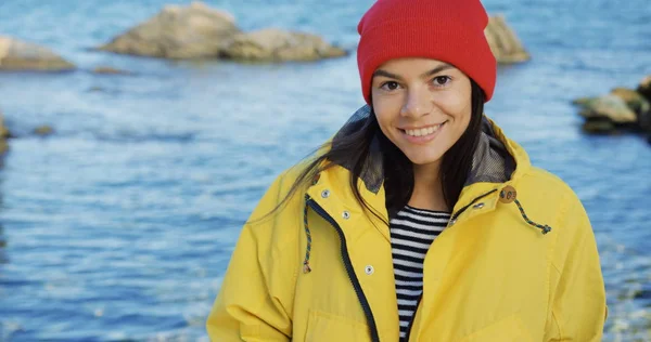 Close up of beautiful young woman in red hat and yellow coat looking on the side and then smiling into the camera on the rocky sea background. Cold sunny day. Portrait shot. Outside.