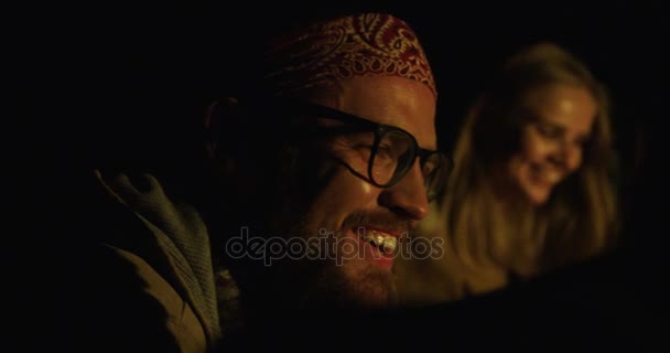 Primer plano del hombre hipster en gafas riendo mientras estaba sentado entre sus amigos cerca del fuego en la noche. Retrato. Al aire libre — Vídeos de Stock