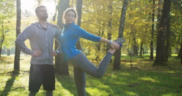 Un paio di corridori che si allungano nel parco soleggiato al mattino presto. Donna che allunga la gamba mentre tiene la spalla dell'uomo. All'aperto — Video Stock