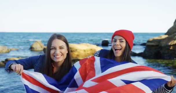 Portrait de deux belles jeunes femmes agitant le drapeau britannique et souriant sur le fond rocheux de la mer. Journée froide mais ensoleillée. Extérieur — Video