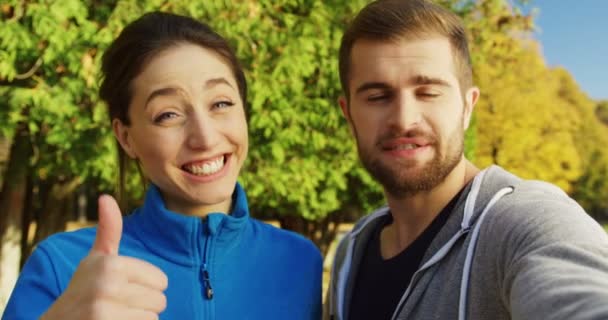 Primer plano de joven sonriente pareja deportiva videochat y mostrando sus pulgares hacia arriba en el soleado parque a principios de otoño. Afuera. Retrato. POV — Vídeos de Stock