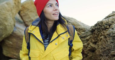 Kırmızı şapka ve kayaların arasında yürüyüş ve etrafa sarı ceket çekici genç kadın. Soğuk güneşli gün. Açık